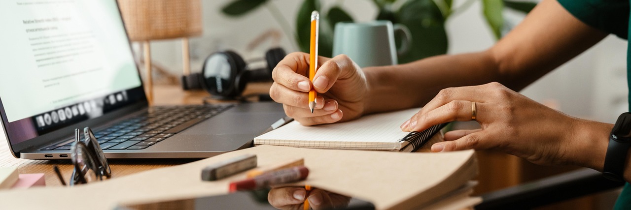 A business owner writes a list of customer questions in a notebook beside an open laptop on a wooden desk, with a mug and headphones behind