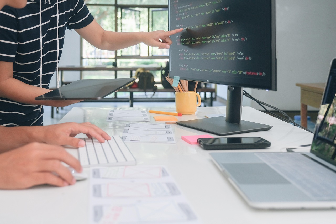 A web developer points at page code on a monitor while a colleague types, with printed page wireframes on a bright office desk